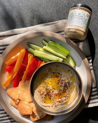Bowl of miso hummus on a plate of sliced vegetables and crackers with a jar of Shared Cultures miso in the corner