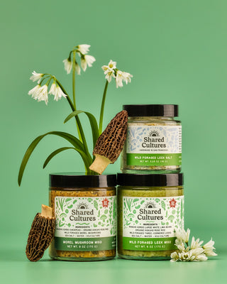 Three jars of Shared Cultures products stacked with decorative morel mushrooms and wild foraged leeks
