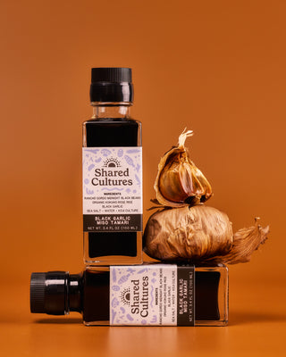Two bottles of Shared Cultures Black Garlic Miso Tamari stacked with two bulbs of black garlic