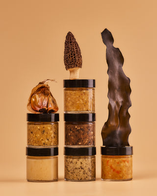Six flavors of Shared Cultures miso taster jars stacked with black garlic cloves, a morel mushroom and piece of kombu